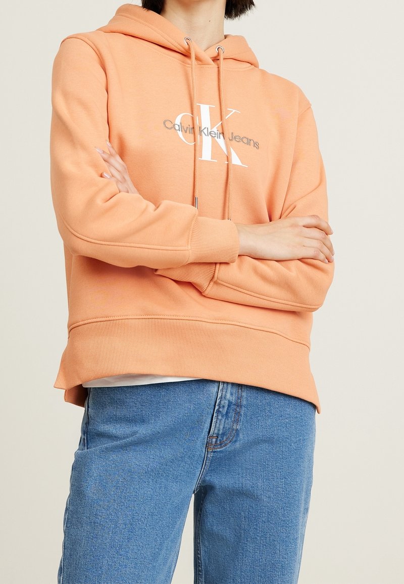 Peach-colored hoodie with a large white "CK" logo and drawstrings. Ribbed cuffs and hem, with a relaxed fit and soft texture. Worn with blue jeans.