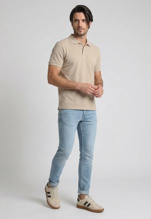 Mand, der står iført en beige poloshirt, lyseblå jeans og beige sneakers med sorte striber mod en ensfarvet baggrund.