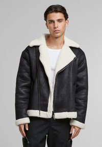 Chaqueta de borreguillo negra con un cuello y puños de color crema, suaves al tacto. Presenta un cierre frontal de cremallera y un diseño de corte relajado.