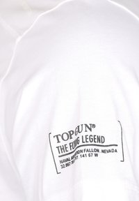 Vit bomulls t-shirt med en svart tryckt etikett på vänstra ärmen, som presenterar "TOP GUN THE FLYING LEGEND" och platsdetaljer.
