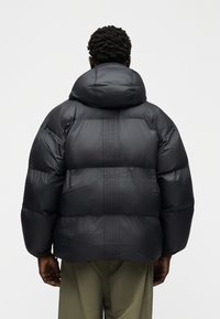 Άνδρας με σκούρο δέρμα, που φοράει μαύρο μπουφάν με κουκούλα, στέκεται γυρισμένος πλάτη μπροστά σε ένα απλό φωτεινό φόντο.