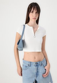 Witte geribbelde crop top met een ritsenkraag, gecombineerd met lichtblauwe high-waisted denim shorts en een kleine denim schoudertas.