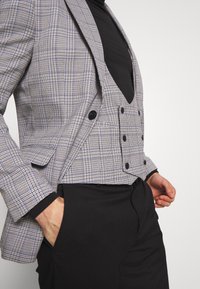 Gepunkteter Blazer in Hellgrau und Schwarz, mit Schalkragen, einer Einzelknopfschließe und zwei Taschen. Über einem schwarzen Rollkragenpullover getragen.