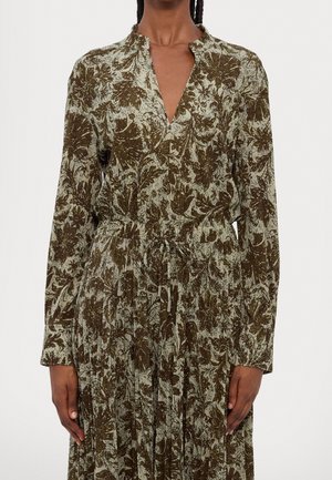 Mujer con vestido de manga larga y escote en V con estampado floral verde y marrón y cintura con cordón, de pie frente a un fondo liso.