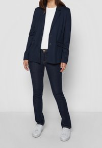 Blazer azul marino con dos botones, solapas con muesca y bolsillos con solapa, combinado con jeans oscuros y zapatillas blancas. Tejido suave y ajuste entallado.