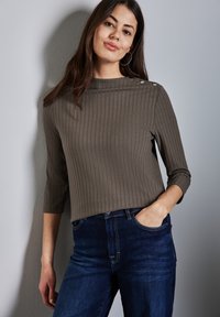 Rippendes, olivgrünes Oberteil mit Dreiviertelärmeln und dekorativen silbernen Knöpfen an der Schulter, kombiniert mit dunkelblauen Jeans.