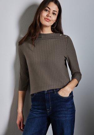 Top a coste verde oliva con maniche a tre quarti e bottoni decorativi argento sulla spalla, abbinato a jeans blu scuro.