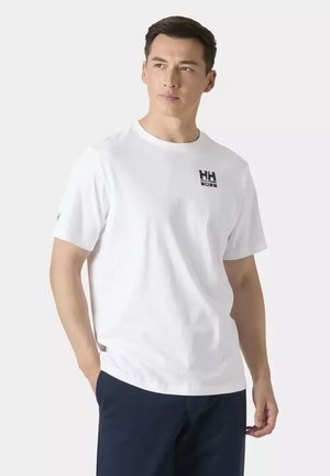 Uomo che indossa una T-shirt bianca Helly Hansen e pantaloni scuri, in piedi con una mano in tasca, guarda di lato contro uno sfondo uniforme.