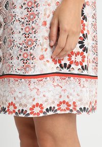 Robe en tissu blanc avec un motif floral en nuances de rouge, noir et gris. L'ourlet présente une bande d'accent contrastante en noir et rouge.