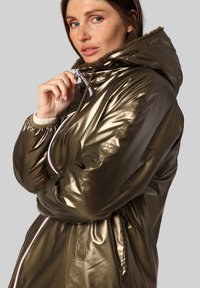 Veste rembourrée vert olive métallique avec une capuche en fourrure, dotée d'une fermeture éclair et d'accents rayés contrastés sur le devant.