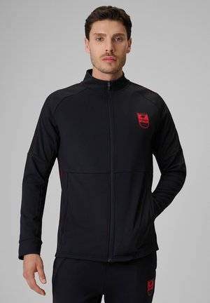 Mężczyzna w czarnej sportowej kurtce zapinanej na zamek z czerwonym logo i dopasowanych czarnych spodniach, stojący na jednolitym szarym tle.