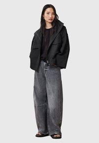 Giacca nera oversize con zip, caratterizzata da ampie tasche, indossata sopra un top scuro e abbinata a jeans a gamba larga grigi chiari e sandali.