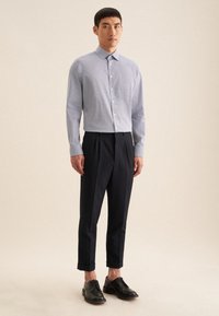 Lichtblauw shirt met patroon en een kraag, gecombineerd met donkere, op maat gemaakte broeken. Het model draagt zwarte leren schoenen. Eenvoudige achtergrond.