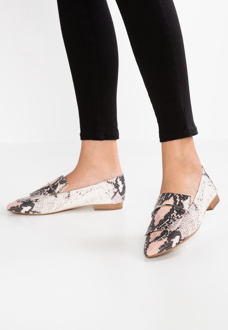 Pieds portant des mocassins à bout pointu et talon bas avec un motif en peau de serpent rose et noire et un détail métallique sur le dessus, associés à un pantalon noir.