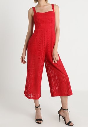 Combinaison en crochet rouge avec encolure carrée sans manches, jambes larges et cropped, et texture à motif subtil. Associée à des talons noirs à bride au niveau de la cheville.