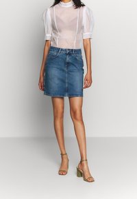 Žena v bílém průhledném topu s volnými rukávy, modré džínsové mini sukni a olivově zelených sandálech na blokovém podpatku s kotníkovým páskem stojí na jednobarevném pozadí.