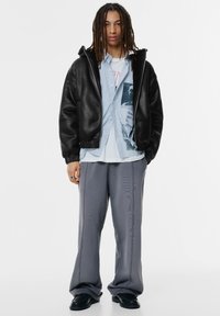 Svart bomberjacka i konstläder, blå randig skjorta över grafisk t-shirt, gråa vida byxor, svarta skor, med elastiska mudd och midjeband.