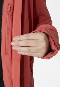 Koralová bunda s hladkým matným povrchem. Má zip detail podél přední části a těsné manžety. Ruka umístěná blízko zipu.