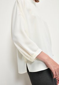 Blusa bianca con maniche lunghe e morbide, con polsini elasticizzati. Presenta un collo alto e fessure laterali all'orlo. Tessuto morbido e leggero.