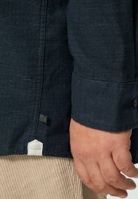 Navyfarbendes Hemd mit Knöpfen, aus strukturiertem Stoff, sichtbarer Naht und weißem Etikett am unteren Ende. Eine Hand ist zu sehen, die die Ärmel des Hemdes trägt.
