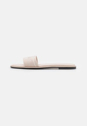 Beige slidesandal med en bred stofrem og flad sål, minimalistisk design på en ensfarvet lys grå baggrund.