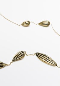 Gouden ketting met verlengde, geribbelde, druppelvormige hangers verbonden door delicate kettingen, met een textuurdetail.