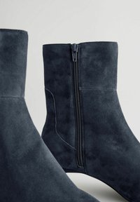 Bottes de cheville en daim gris foncé avec une fermeture éclair sur le côté, texture lisse et silhouette élégante. Le dessus est légèrement structuré avec des coutures minimales.