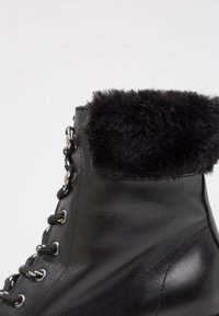 Botín de cuero negro con un acogedor cuello de piel de fur negro, ojales metálicos y cordones negros, con un acabado suave y brillante en el cuerpo.