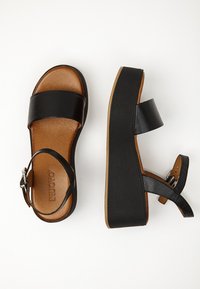 Černé kožené platformové sandály s širokým předním páskem, nártní páskem se sponou a hnědou polstrovanou stélkou na bíl�ém pozadí.