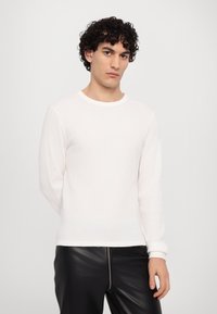 Hvid ribbet sweater med lange ærmer og rund halsudskæring, lavet af struktureret stof, parret med sorte bukser i imiteret læder, stående mod en ensfarvet baggrund.