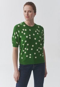 Pull en tricot vert à manches courtes, avec des pois blancs sur l'ensemble et un col rond. Texture douce et design ajusté.
