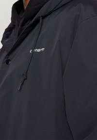 Svart huva jacka med dragsko och vit Carhartt-logotyp broderad på bröstet, bärs över en svart hoodie.