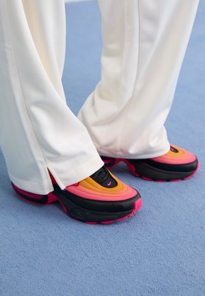 Pieds portant des baskets dégradées orange, rose et noire avec un pantalon blanc, debout sur un sol recouvert de moquette bleue.