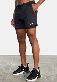 Shorts de bain noirs avec une ceinture élastique et un cordon de serrage, arborant un logo blanc sur la jambe gauche, assortis à des chaussures de sport noires.