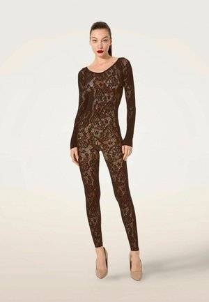 Woman wearing black lace fitted bodysuit and beige high heels standing against a plain light background.