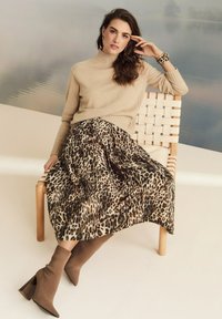 Vrouw die zit op een geweven houten stoel, gekleed in een beige coltrui, een rok met luipaardprint, een bijpassende armband en bruine hakken.