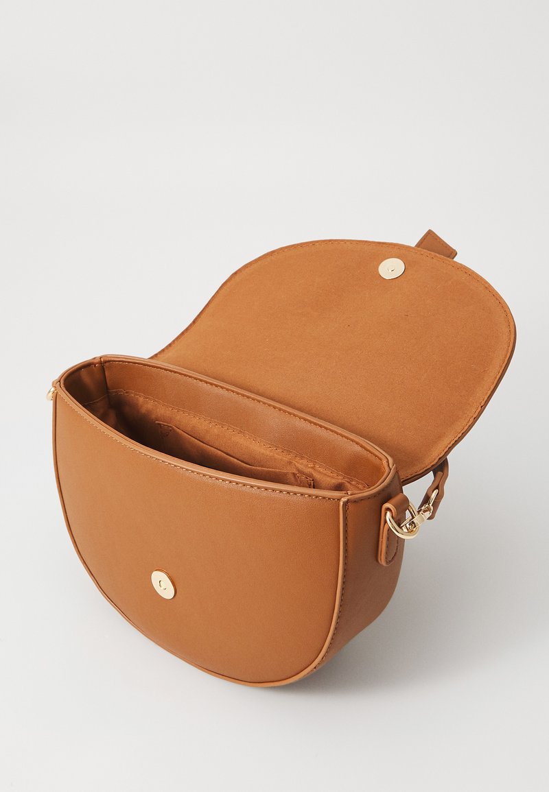 Sac en cuir tan à bandoulière avec une forme arrondie, fermeture magnétique, et un intérieur doux. Il dispose d'un seul compartiment et de quincaillerie dorée.