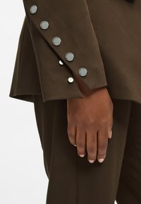 Brauner Blazerärmel mit fünf silbernen Druckknöpfen und einem glatten, strukturierten Stoff. Hand auf dem Blazer platziert.