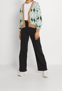 Sweter w romby w zielono-białym kolorze z zapięciem na guziki i brązowym obszyciem, noszony na białej bluzce i czarnych spodniach, zestawiony z jasnymi trampkami.