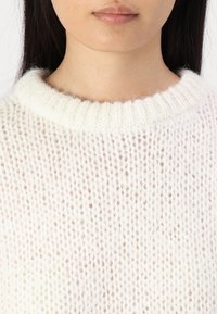 White knitted sweater with a ribbed crew neckline, featuring a loose, airy texture and a subtle gradient effect towards the bottom.