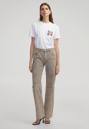 T-shirt blanc à manches courtes avec une poche décorative, associé à un jean évasé beige à motifs. Des sandales à talons hauts complètent le look.