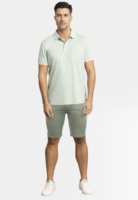 Polo verde claro con mangas cortas, un cuello y un pequeño bolsillo en el pecho. Emparejado con pantalones cortos a juego y zapatillas blancas.