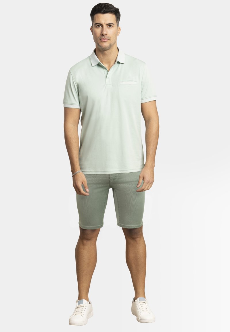 Polo verde claro con mangas cortas, un cuello y un pequeño bolsillo en el pecho. Emparejado con pantalones cortos a juego y zapatillas blancas.