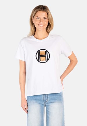 Glimlachende vrouw met blond haar, gekleed in een wit T-shirt met een zwart-gouden geometrisch logo, gecombineerd met lichtblauwe jeans.