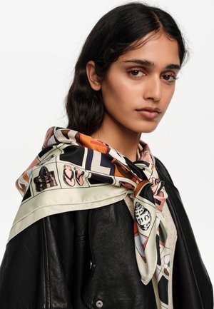 Joven con cabello largo y oscuro que lleva una bufanda de seda con un patrón colorido sobre una chaqueta de cuero negra, mirando ligeramente hacia un lado contra un fondo blanco.