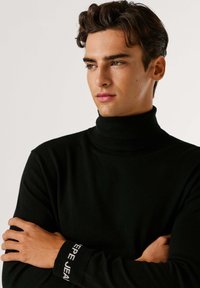 Maglione a collo alto nero realizzato in morbido tessuto, dalla vestibilità attillata, con polsini a costine caratterizzati da testo bianco. Il modello ha i capelli tagliati corti e un'espressione neutra.