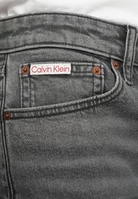 Pelēku džinsu bikses ar maigu tekstūru, kam ir sarkans un balts "Calvin Klein" zīmogs, vara kniedes un klasiskā piecu kabatu dizains.