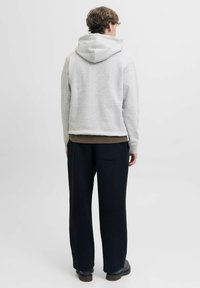 Mężczyzna stojący tyłem, ubrany w jasnoszary hoodie, ciemne szerokie spodnie oraz ciemne buty na jednolitym jasnym tle.