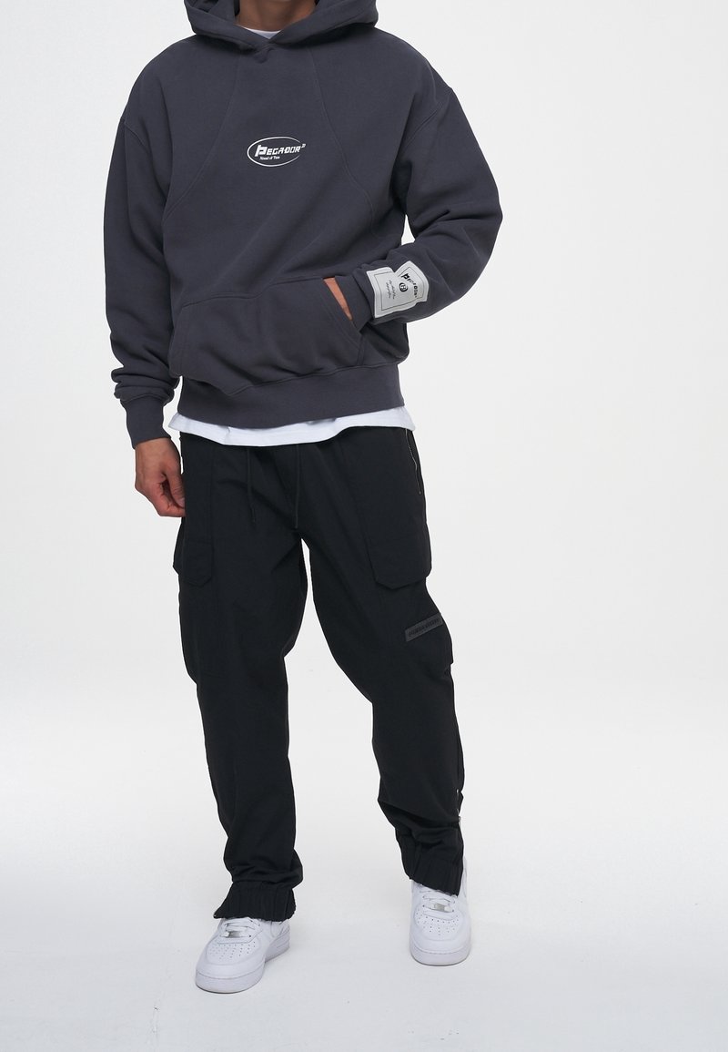 Felpa grigia con cappuccio e tasca frontale, decorata con logo, abbinata a pantaloni cargo neri con tasche e polsini elasticizzati, scarpe da ginnastica bianche.