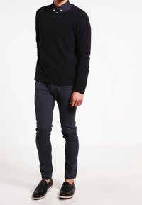 Zwarte gestructureerde trui, donkere denim slim-fit jeans en zwarte schoenen met een witte zool. Gelaagd met een overhemd eronder.